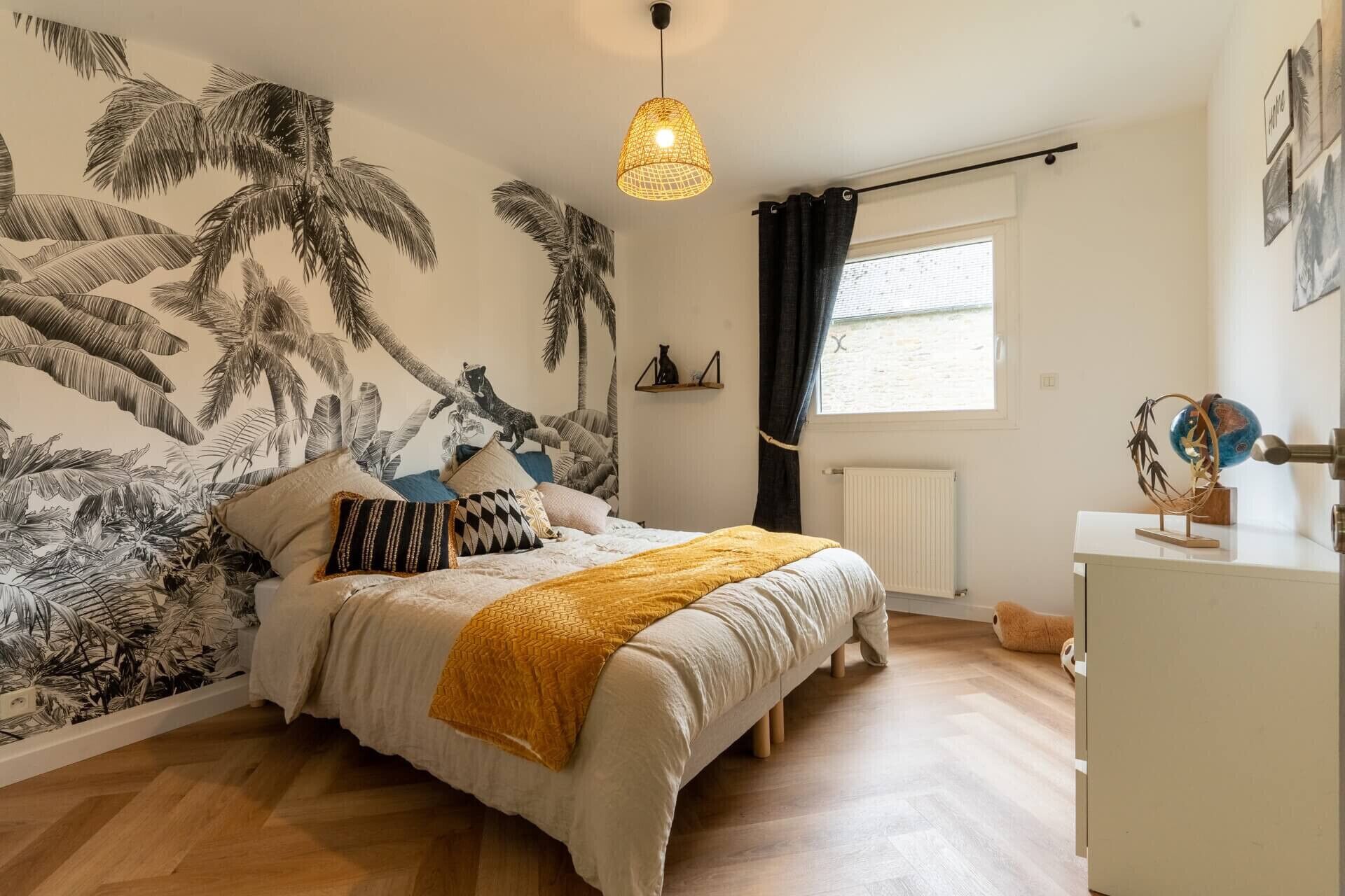 Een slaapkamer met een bed, een hanglamp, een raam met gordijnen en een muur met een tropische muurschildering.