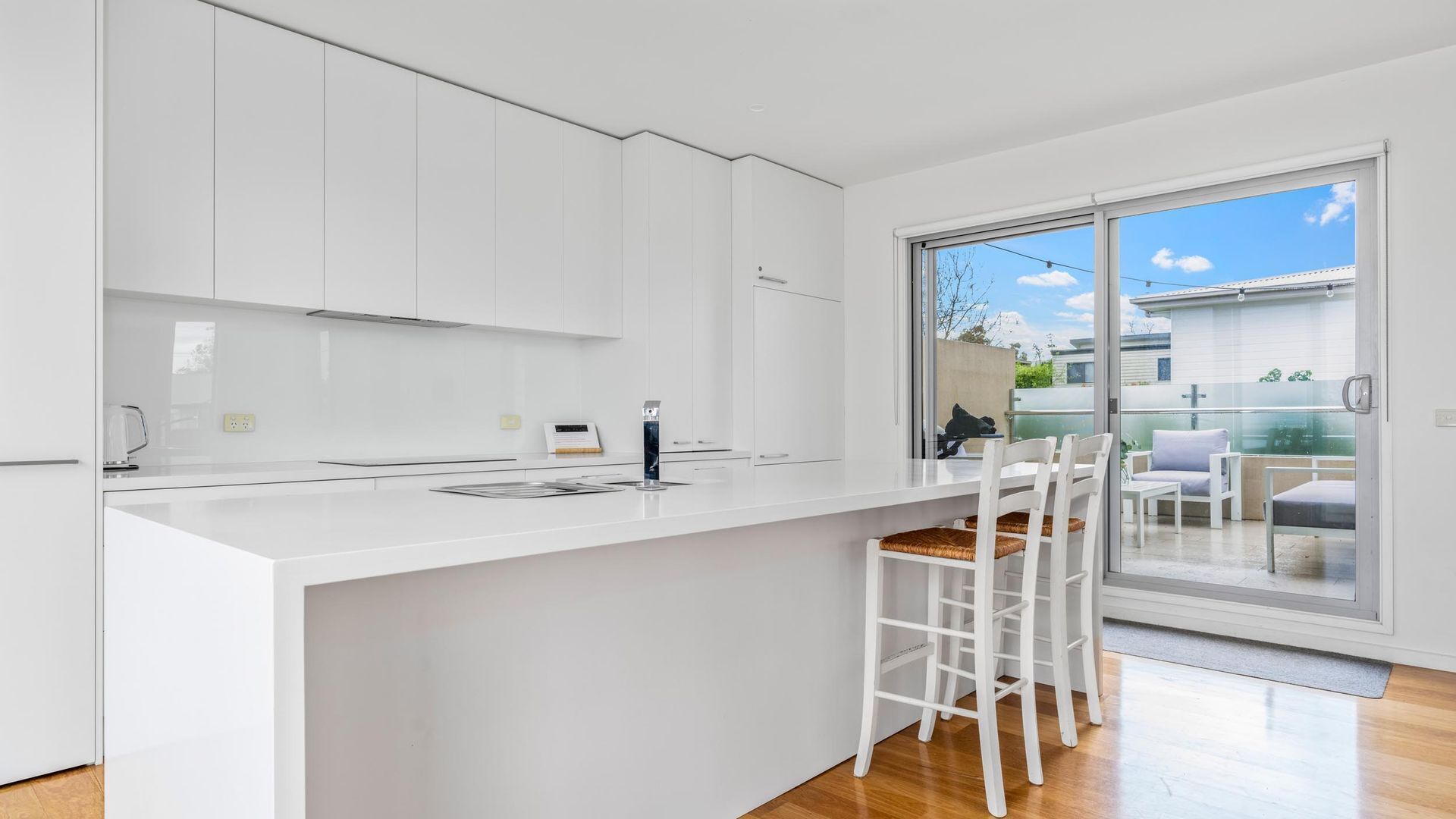 A modern kitchen with white cabinetry, a central island, and a sliding glass door leading to an outdoor seating area.