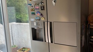 Un réfrigérateur Samsung en acier inoxydable, doté d’un distributeur d’eau et orné de divers aimants et autocollants, se trouve dans une cuisine avec une porte-fenêtre coulissante donnant sur un balcon où sont disposées des plantes en pot.