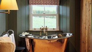 A bathroom with a freestanding bathtub, a chair, a window with a view of greenery, and a wooden cabinet. - JOIVY Lathallan Mill Farmhouse (Leven)