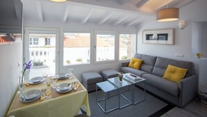 Una habitación luminosa con una mesa de comedor lista para dos personas, un sofá gris con cojines amarillos y una mesa de centro de vidrio.