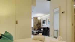 Habitación de hotel con una cama, dos toallas enrolladas, un almohadón verde, una mesita pequeña y una pared de piedra al fondo.