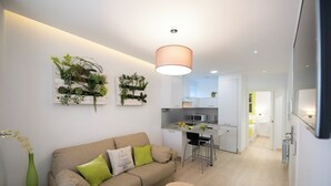 Una sala moderna con un sofá beige, cojines verdes, una pequeña mesa redonda con un vaso y una manzana verde. Se ve una cocina con armarios blancos y un microondas al fondo.