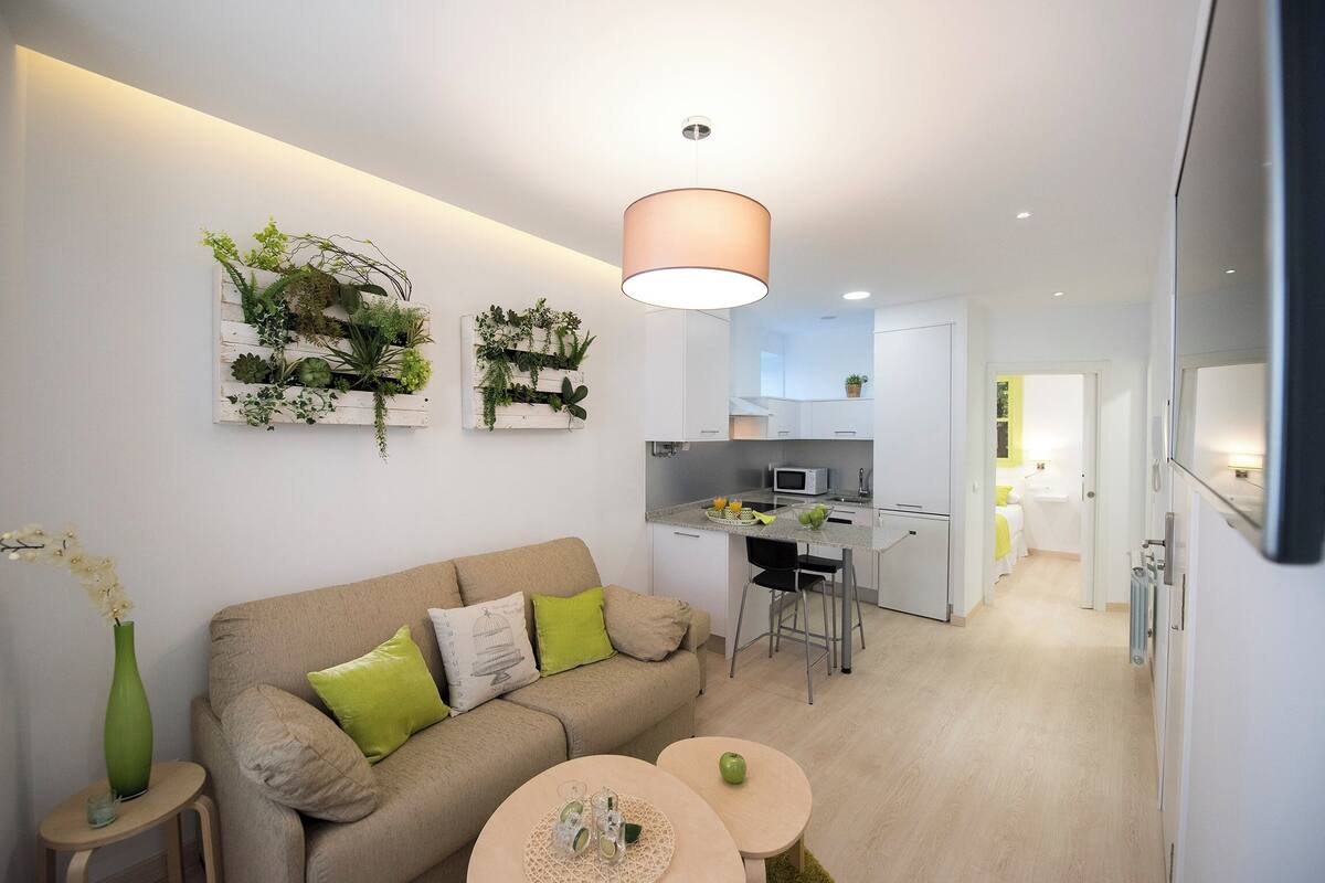 A modern living room with a beige sofa, green cushions, a small round table with a glass, and a green apple. A kitchen with white cabinets and a microwave is visible in the background.