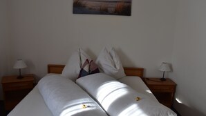 Cama bem arrumada com lençóis brancos, duas mesas de cabeceira com abajures e um quadro de uma paisagem na parede.