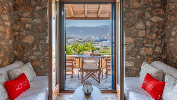 A room with a stone wall, a sofa with red pillows, a table with a candle, and a view of a city and mountains through a glass door. - Elena's Traditional DreamHouse (Paros)