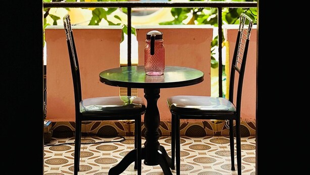 An outdoor seating area with a table and chairs, a view of greenery through an archway, and a tiled floor. - Shalos Inn (Puducherry)