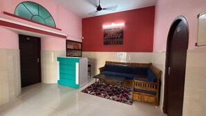 A room with a red wall, a blue sofa, a wooden bench, a turquoise cabinet, and a tiled floor. - Shalos Inn (Puducherry)