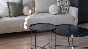 Ein modernes Wohnzimmer mit einem hellen Sofa, zwei runden schwarzen Tischen und einer dunkel gestrichenen Wand.