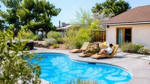 A swimming pool with lounge chairs, surrounded by greenery and a building with a tiled roof.