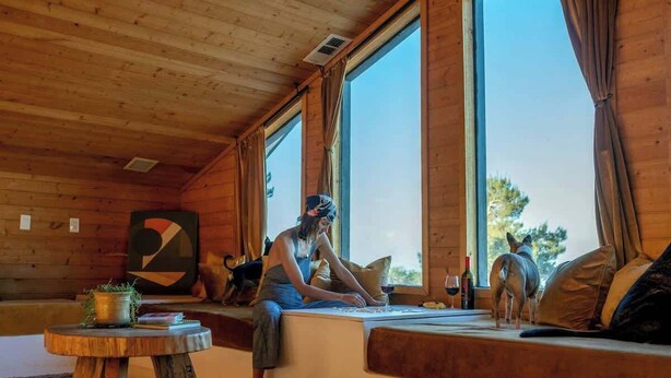 A cozy cabin interior with a large window, wooden walls, a round wooden table, and a dog on the window seat.