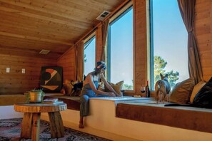 Interior acogedor de una cabaña con un ventanal grande, paredes de madera, una mesa redonda de madera y un perro en el asiento junto a la ventana.