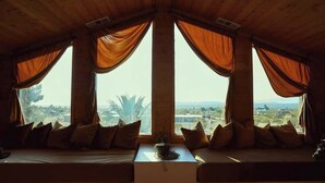 A room with a wooden ceiling, large windows with orange curtains, a sofa with cushions, and a view of palm trees and buildings.