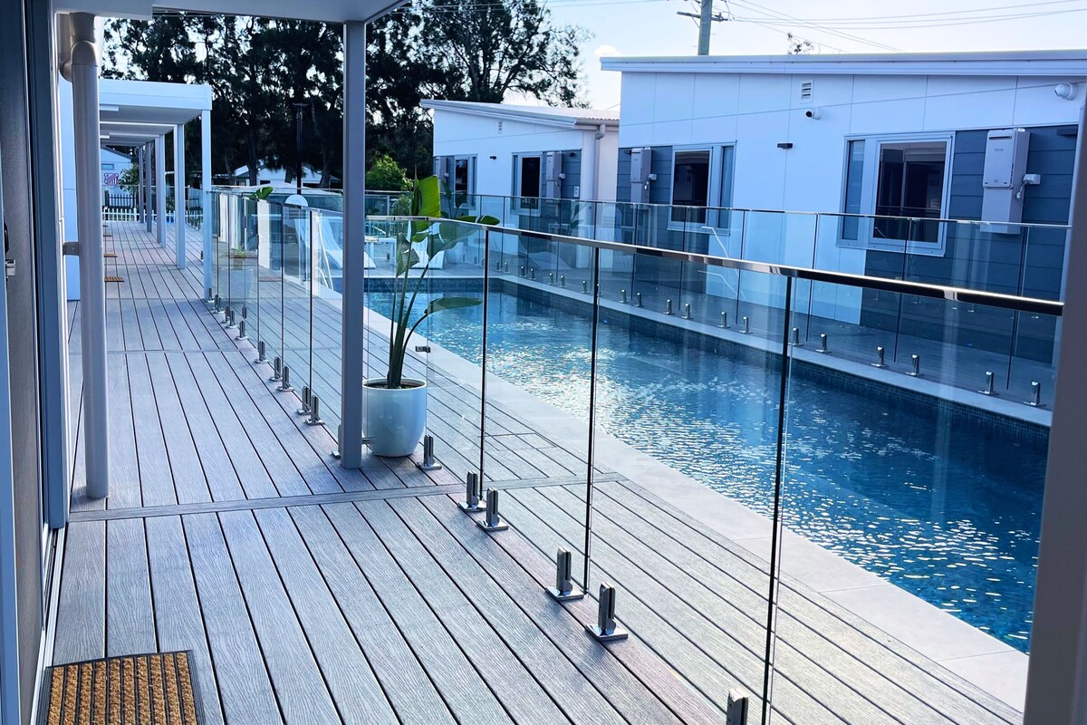A wooden deck with a glass railing overlooking a swimming pool, a covered area, and a building in the background.