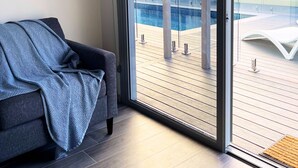 A modern living room with a grey sofa and a large sliding glass door leading to a wooden deck with a pool. - Resort on Cedar (Windang)