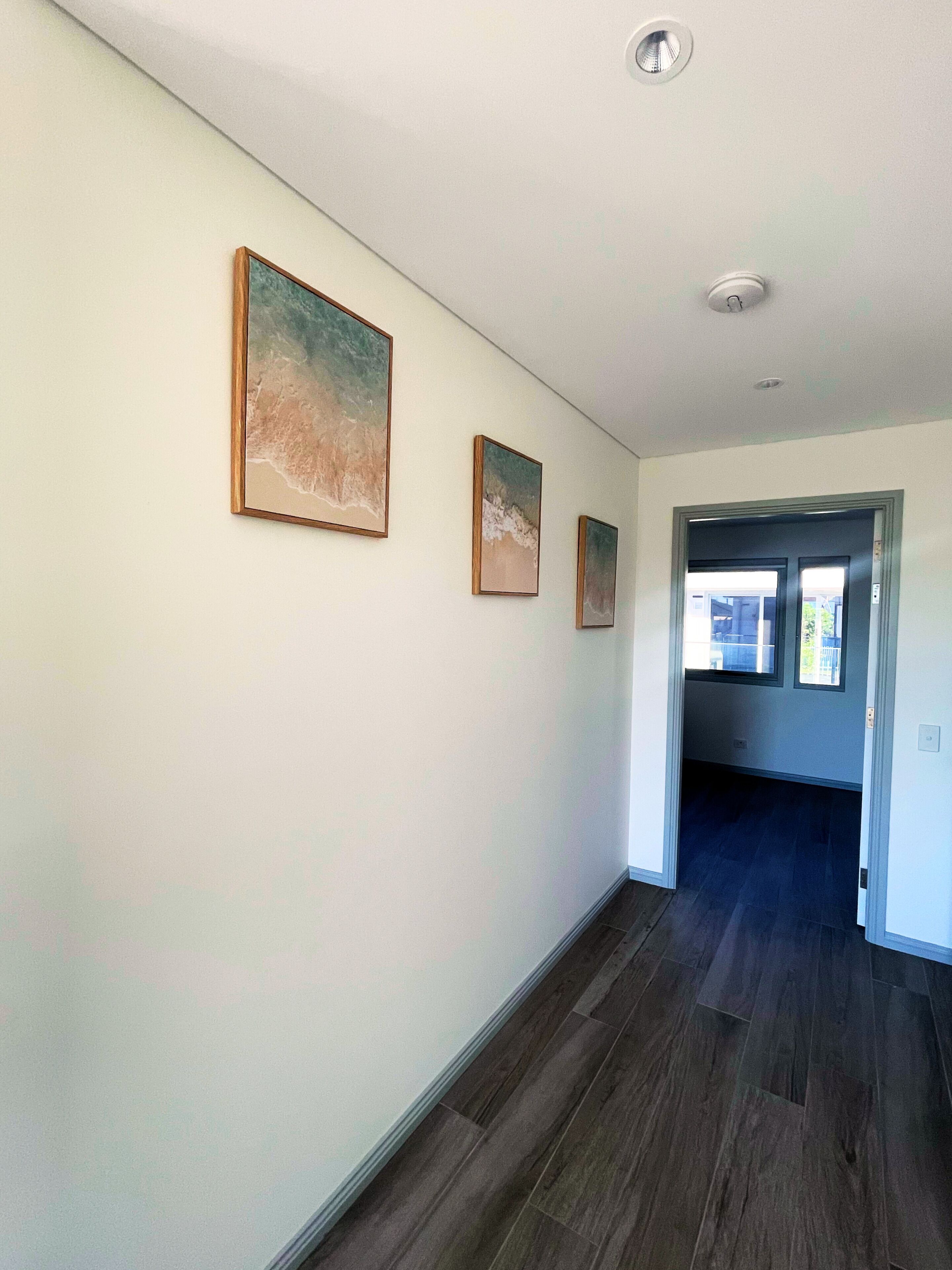 A hallway with three framed artworks on the wall, a wooden floor, and a door leading to another room.