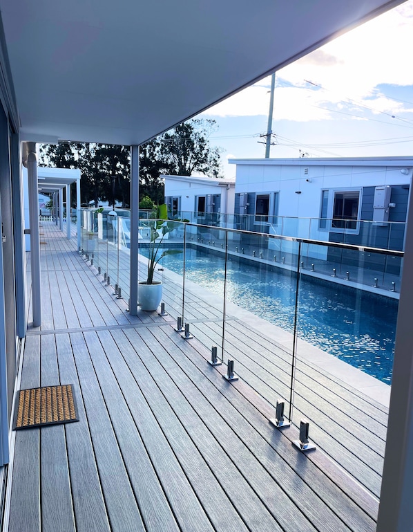 A wooden deck with a glass railing overlooking a swimming pool, a covered area, and a building in the background. - Resort on Cedar (Windang)