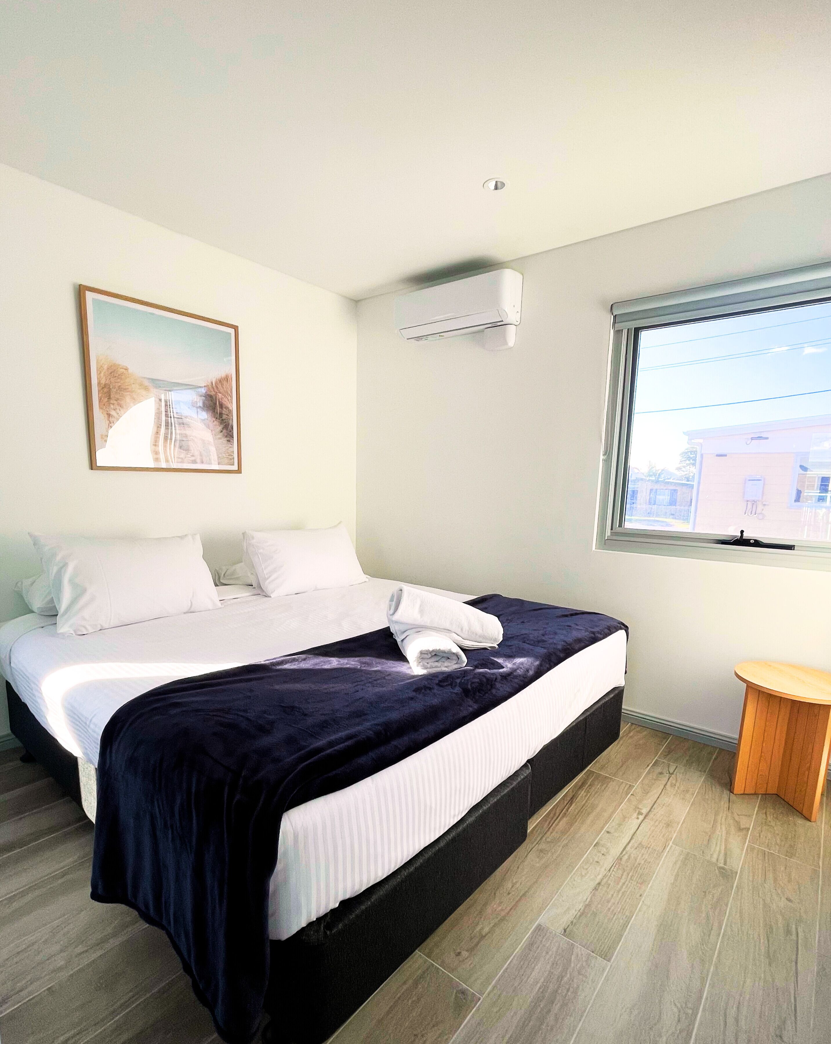 A hotel room with a bed, a small wooden side table, a window with a view, and an air conditioning unit.