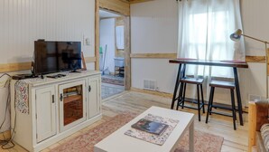 Ein Zimmer mit Fernseher auf einem Schrank, einem kleinen Tisch mit einer Zeitschrift, einem Fenster mit Vorhängen und einem Sessel.