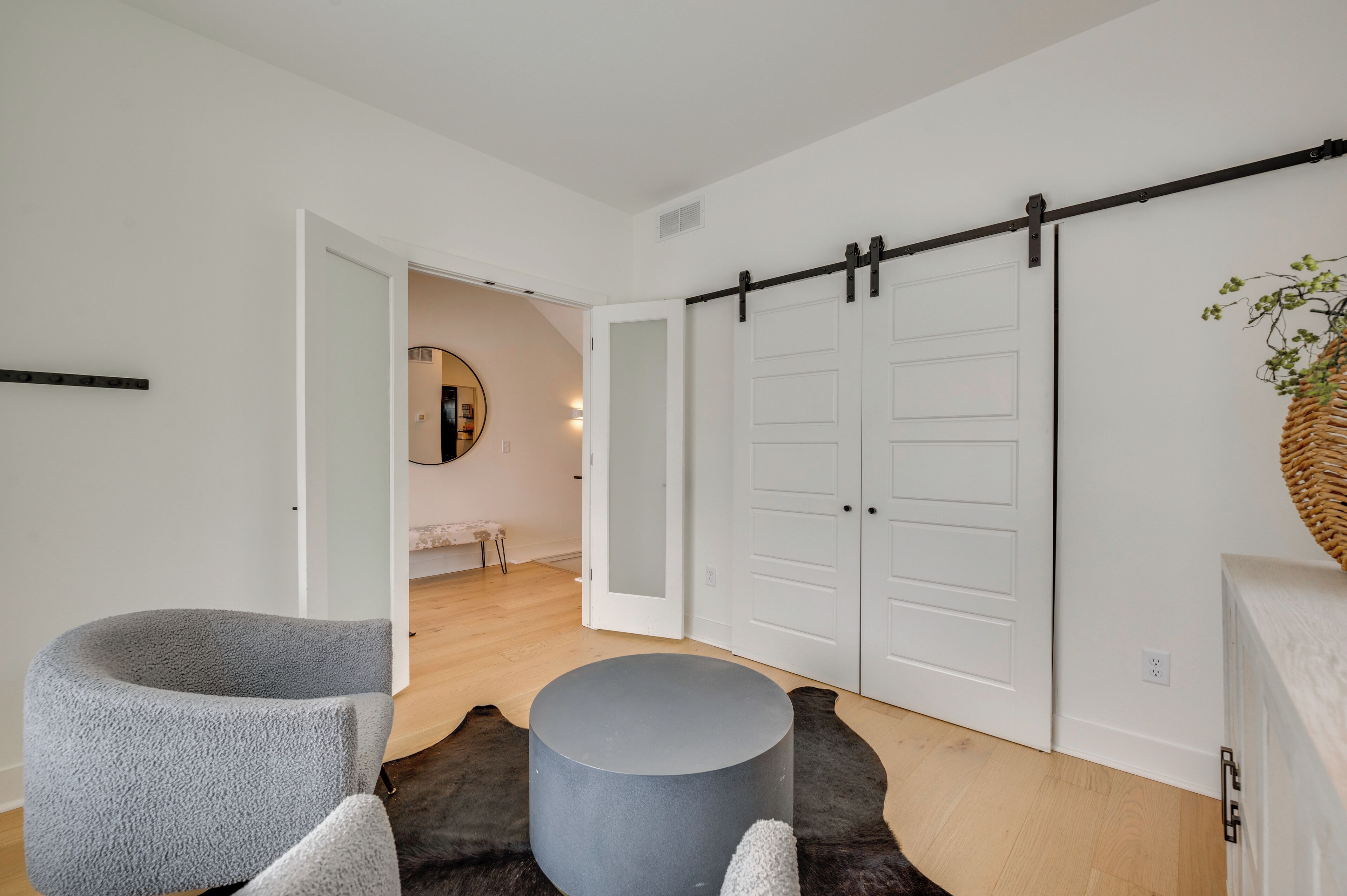 A modern living room with a grey armchair, a round ottoman, and a large mirror on the wall. There is a sliding barn door closet and a wooden floor.