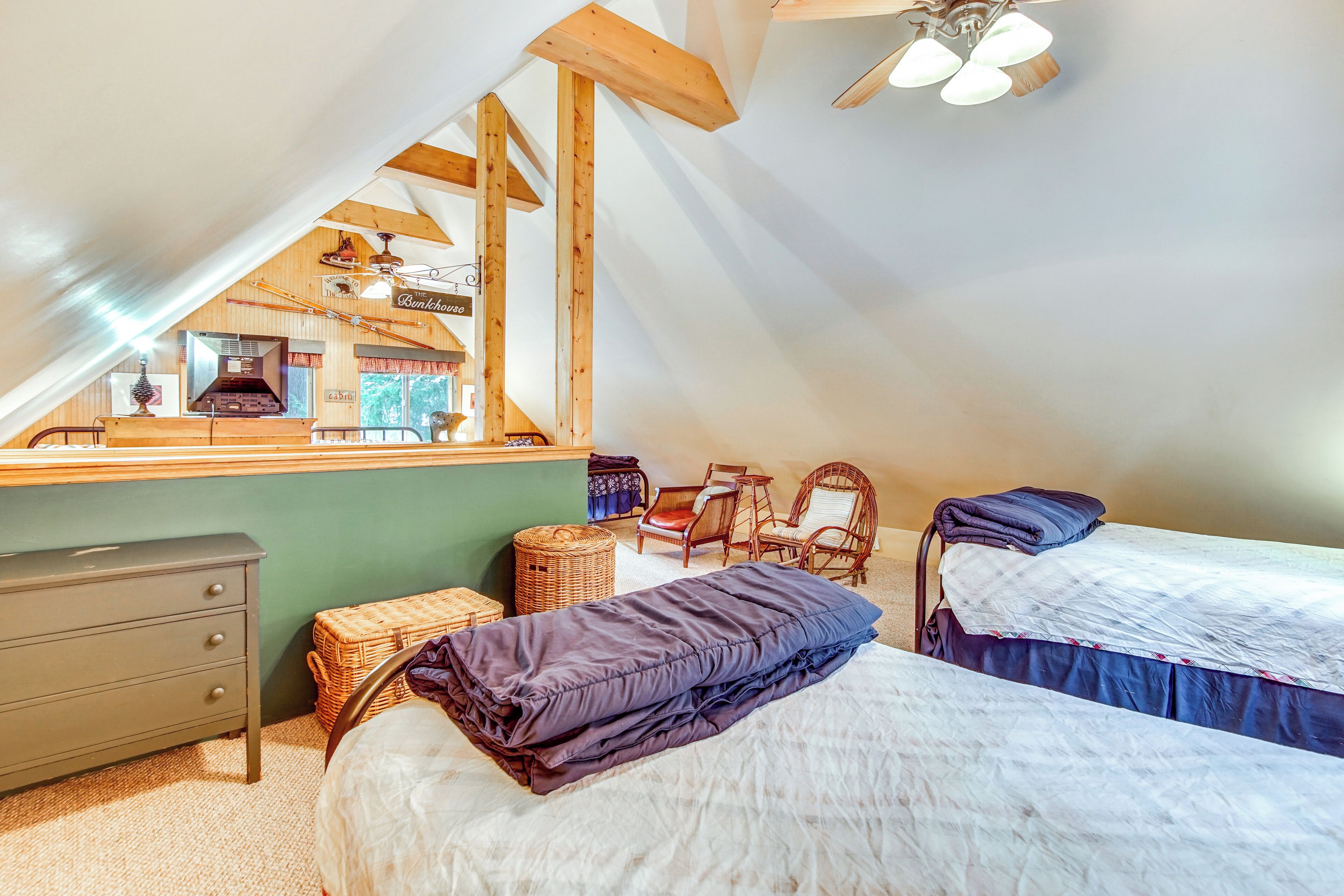 A cozy attic bedroom with two beds, a dresser, wicker baskets, and a view of the kitchen.