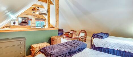 A cozy attic bedroom with two beds, a dresser, wicker baskets, and a view of the kitchen.