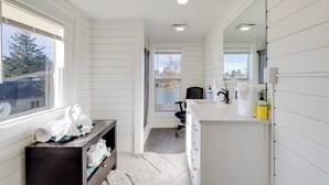 A modern bathroom with white walls, a large window, a vanity with a mirror, and a chair. - Walk to Beach: Riverside Retreat in Seaside! (Seaside)