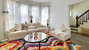 A living room with two white sofas, a glass coffee table, and a colorful rug. There are curtains on the windows, a lamp on a side table, and a staircase in the background. - Walk to Beach: Riverside Retreat in Seaside! (Seaside)