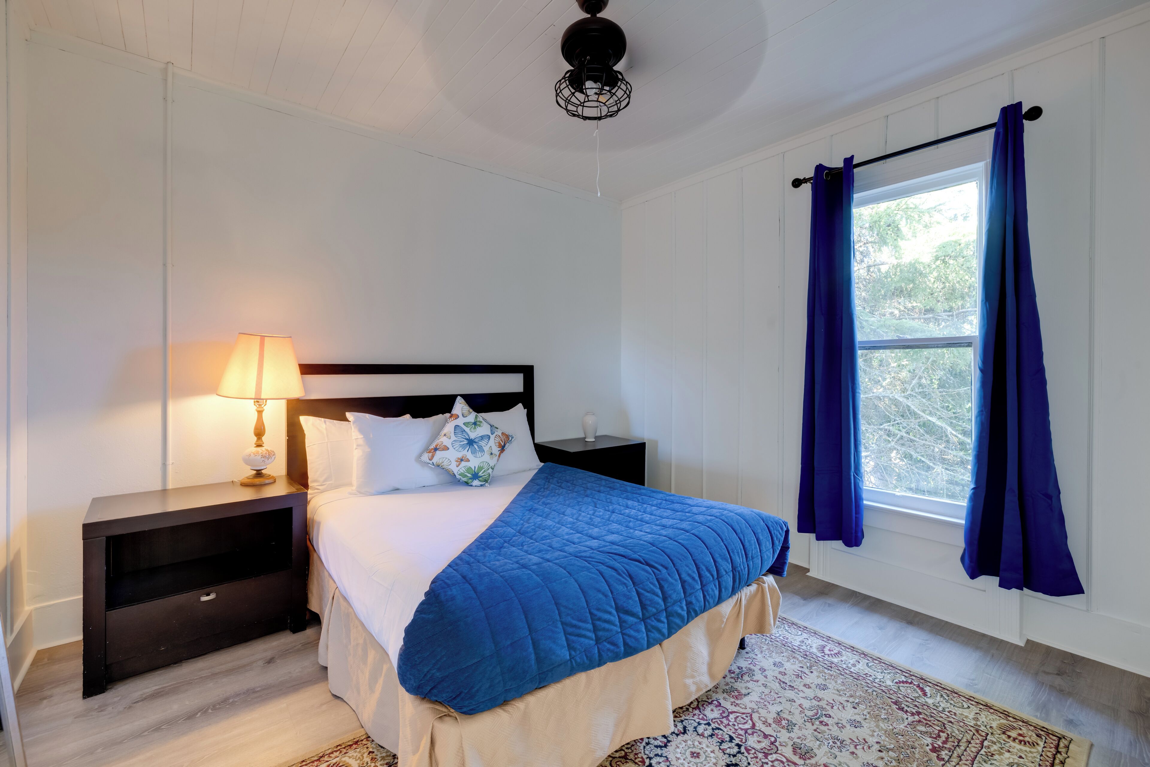 Un dormitorio con cama, mesita de noche, lámpara, ventana con cortinas y alfombra estampada.