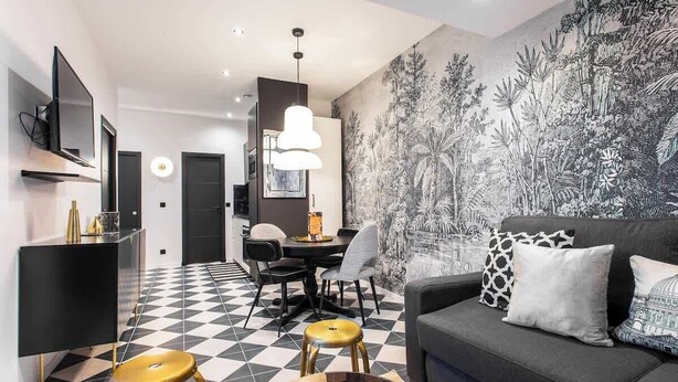 A modern living room with a checkered floor, a grey sofa with patterned cushions, a dining area with chairs and a table, and a wall with a botanical mural.