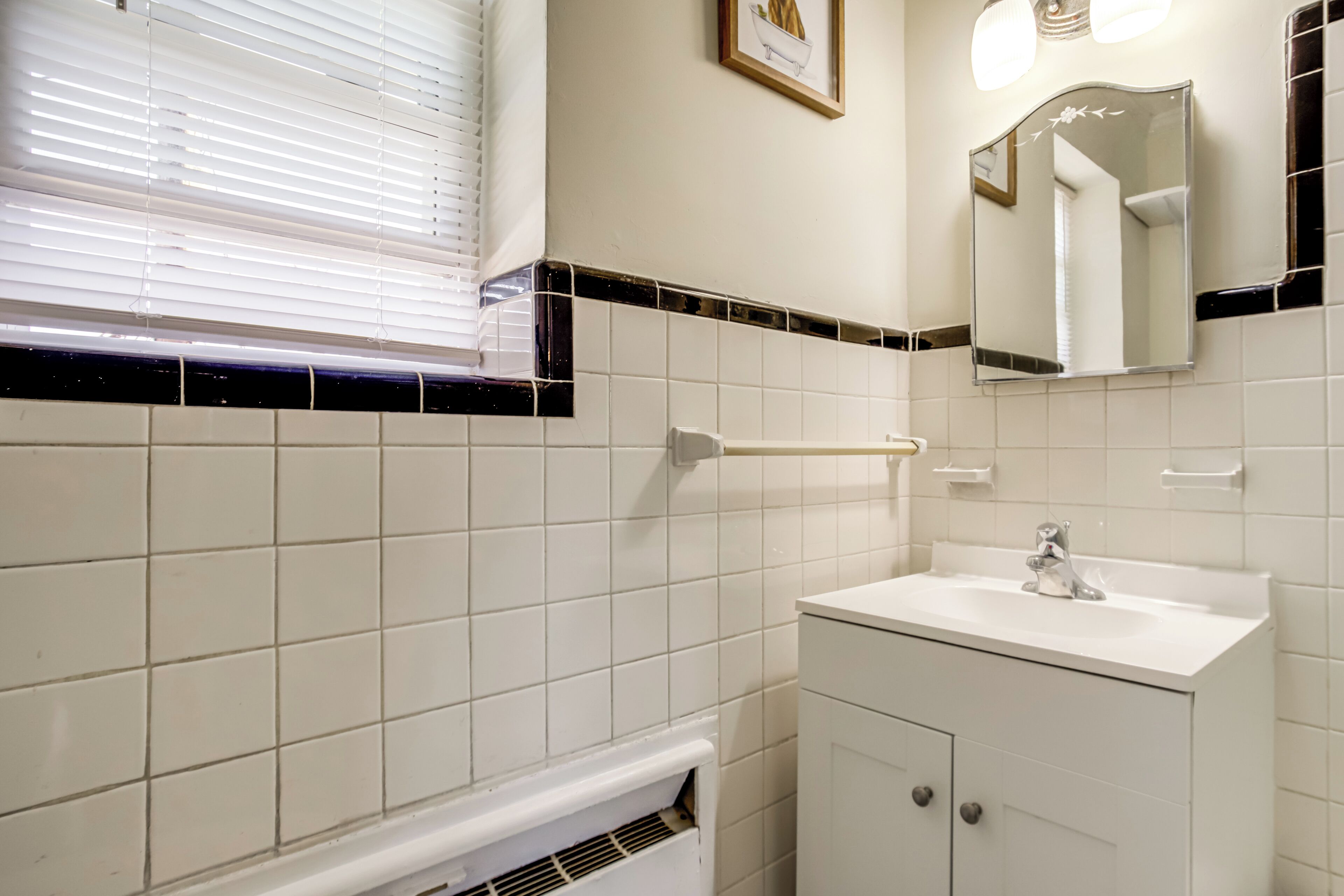 Une salle de bain avec des carreaux blancs, un lavabo blanc, un miroir et une fenêtre avec des stores.