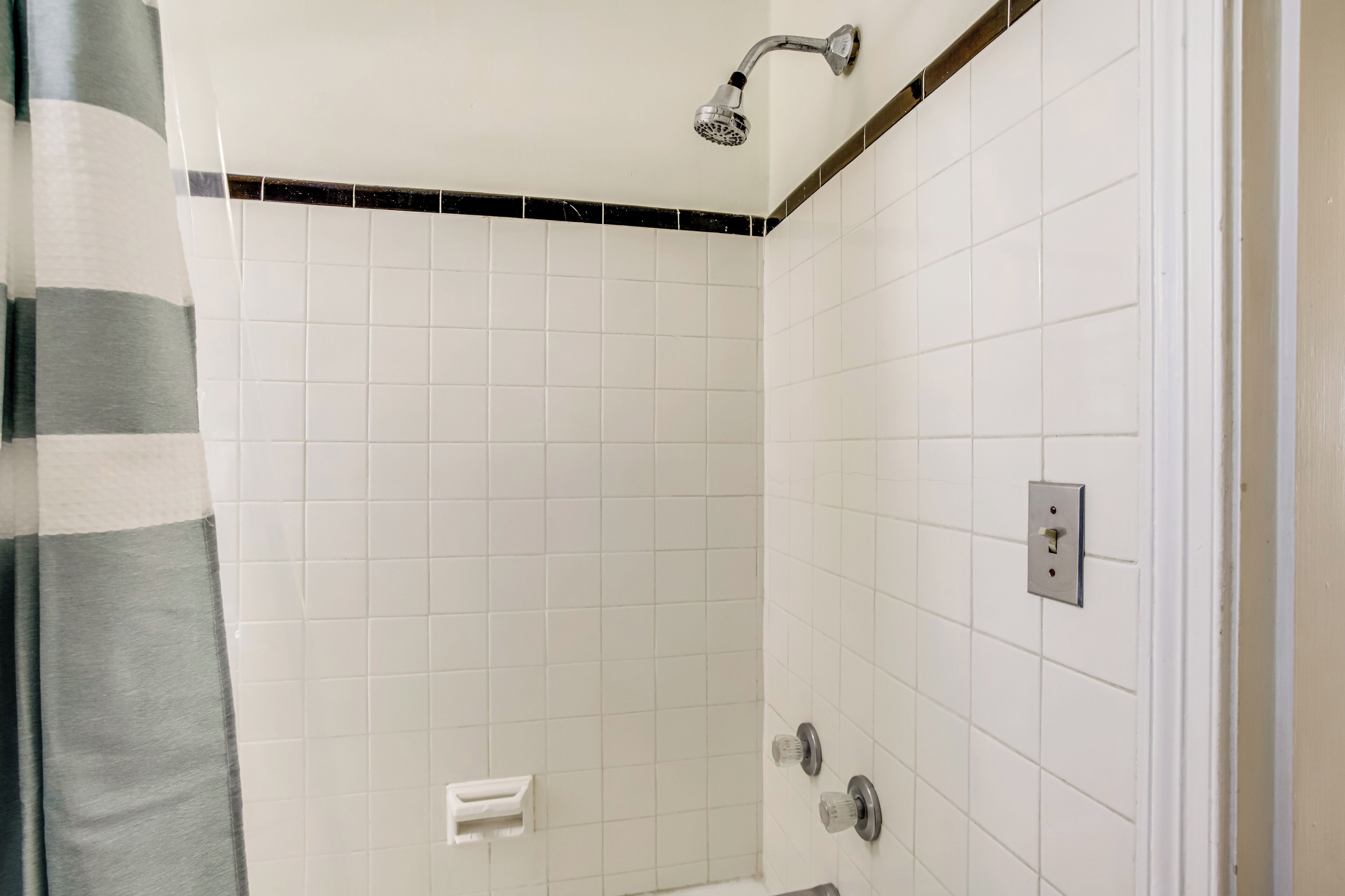 A bathroom with white tiled walls, a showerhead, a towel rack, and a light switch.