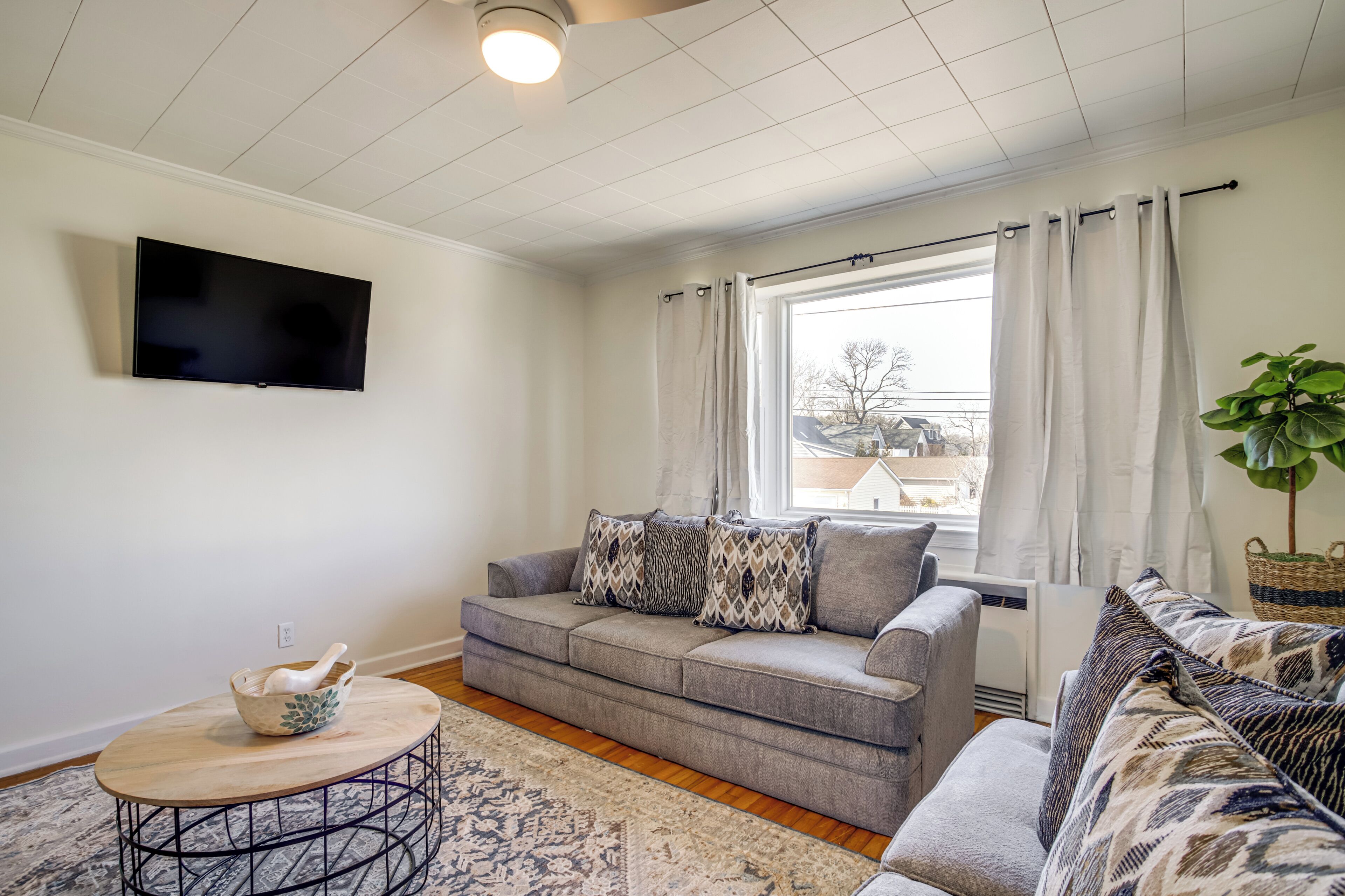 A living room with a grey sofa, a round coffee table, a flat-screen TV mounted on the wall, and a window with curtains.
