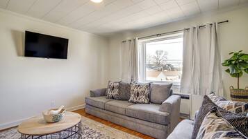 A living room with a grey sofa, a round coffee table, a flat-screen TV mounted on the wall, and a window with curtains.