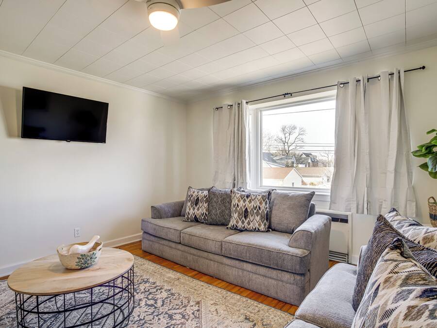 A living room with a grey sofa, a round coffee table, a flat-screen TV mounted on the wall, and a window with curtains.