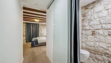 A narrow hallway leading to a bedroom with a stone wall and a wooden ceiling. A bed is visible in the room, and a toilet is seen in the adjacent bathroom.