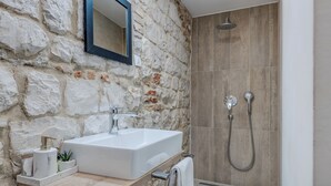 A bathroom with a stone wall, a white sink, a mirror, and a shower area. - The Code Luxury Rooms (Split)