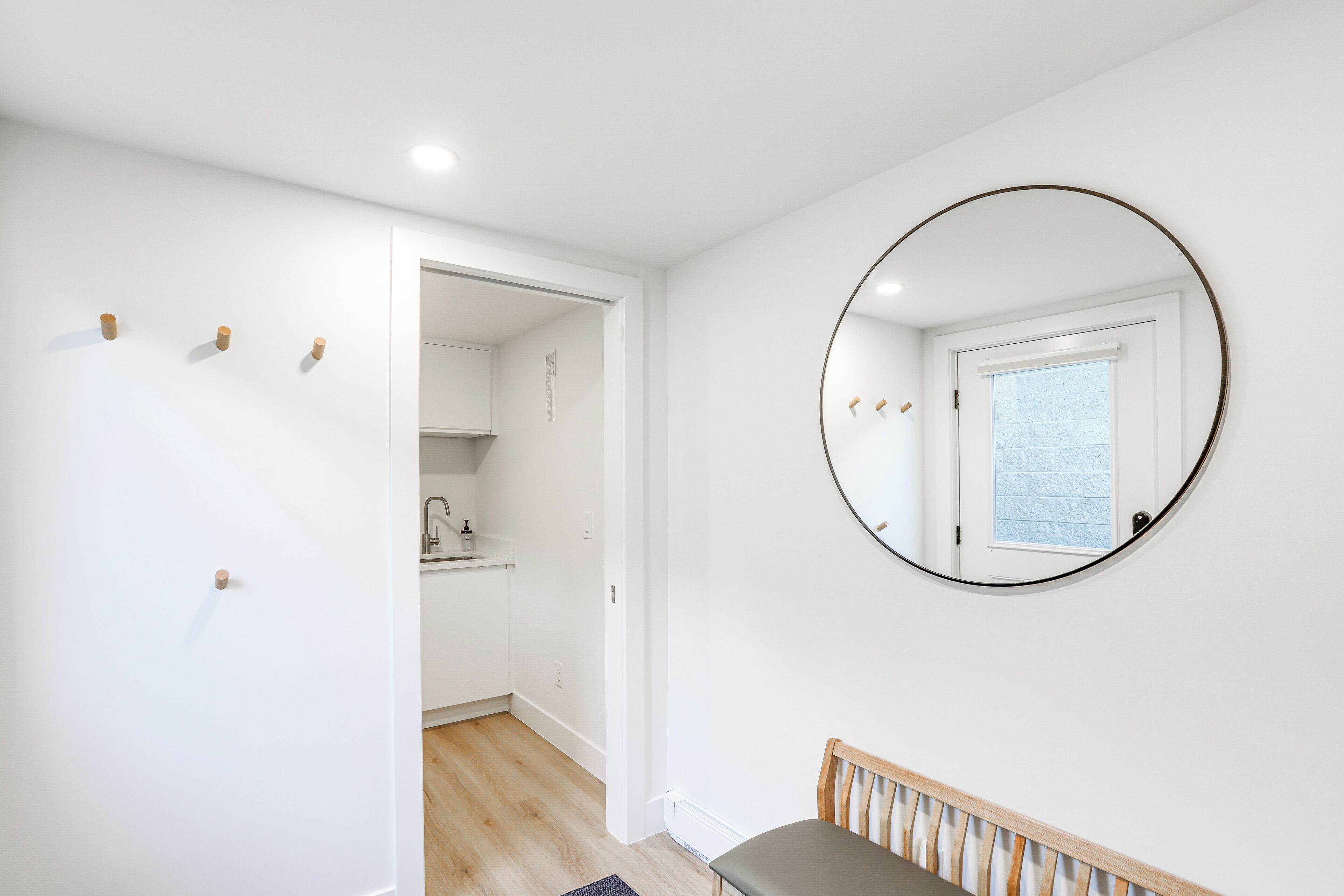 A room with a wooden bench, a round mirror on the wall, and a view of a kitchen through an open door.