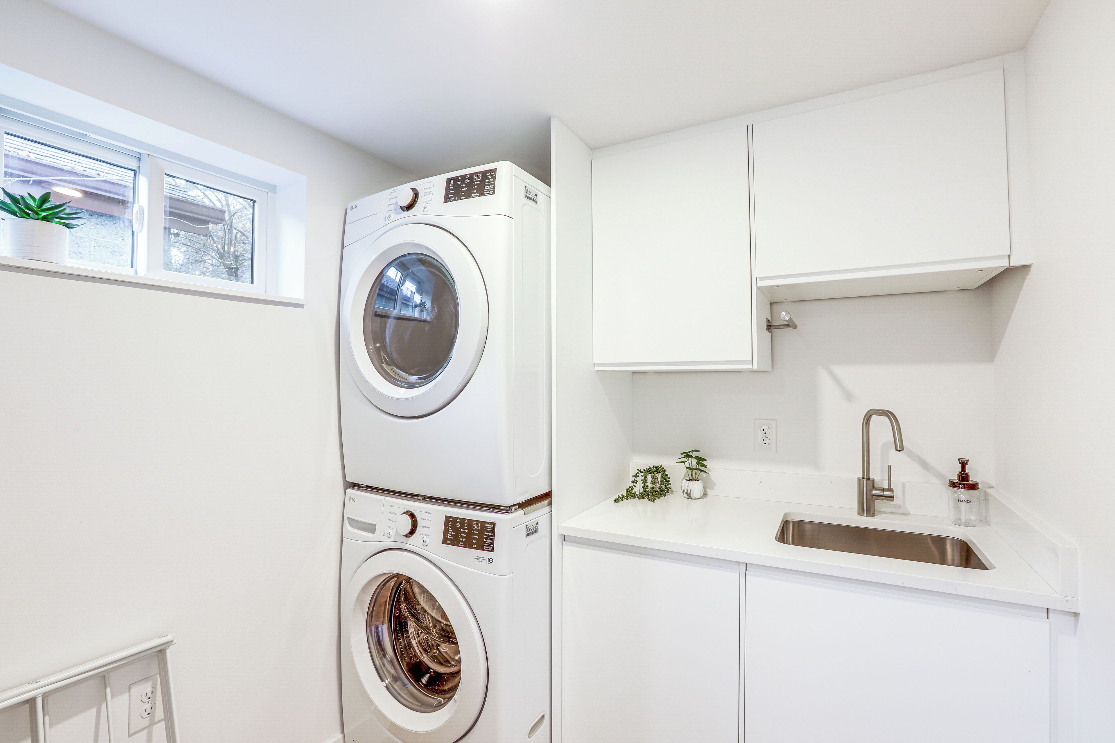 A compact laundry room with a stacked washer and dryer, a sink, and a small window with a plant.