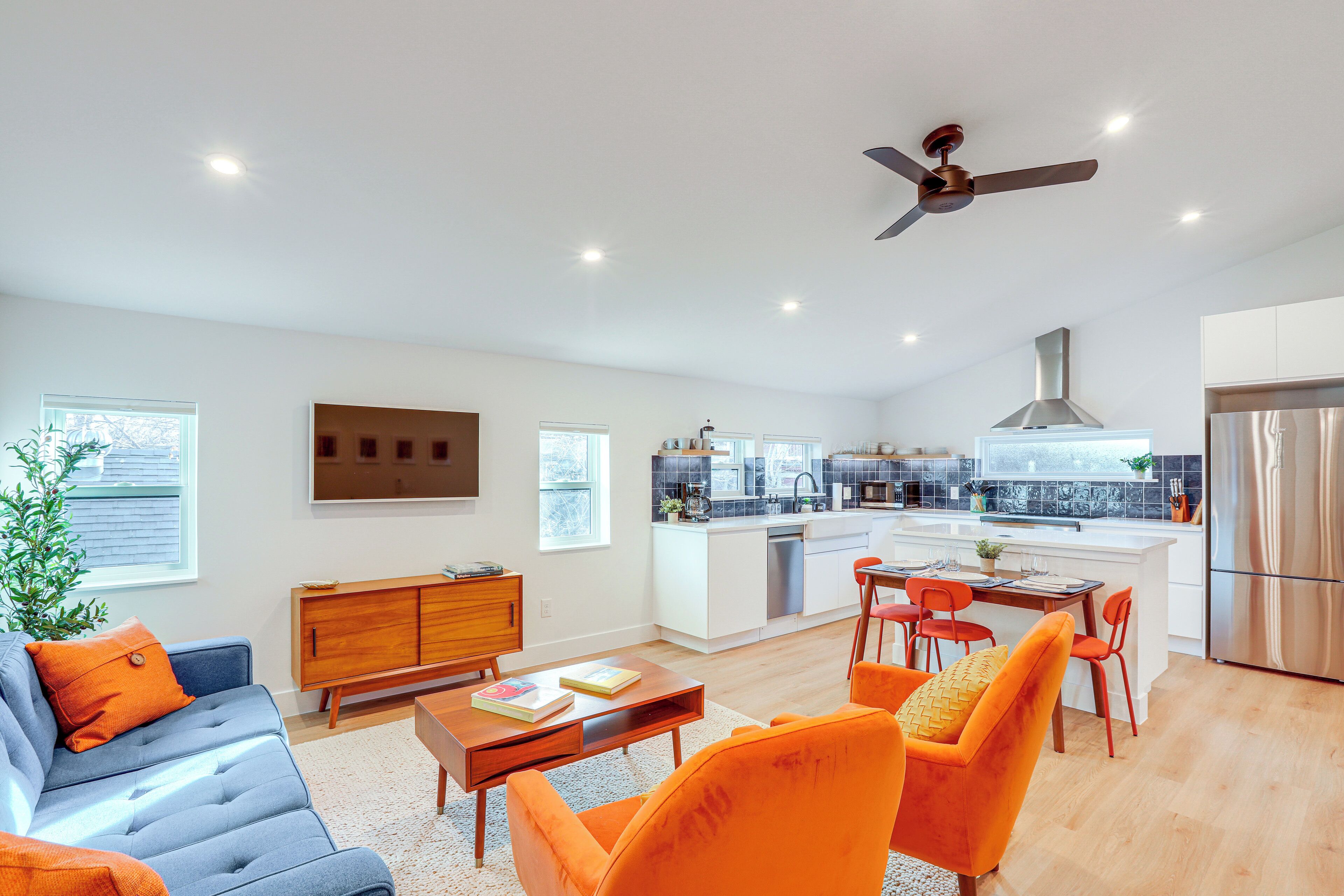 Een moderne woonkamer met een bank, salontafel en oranje stoelen. Achtergrond toont een keuken met witte kasten en roestvrijstalen apparatuur.