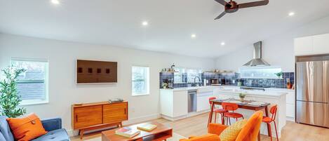 A modern living room with a sofa, coffee table, and orange chairs. A kitchen with white cabinets and stainless steel appliances is visible in the background.
