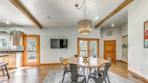 A spacious dining area with a wooden table and chairs, a hanging light fixture, and a television on the wall.