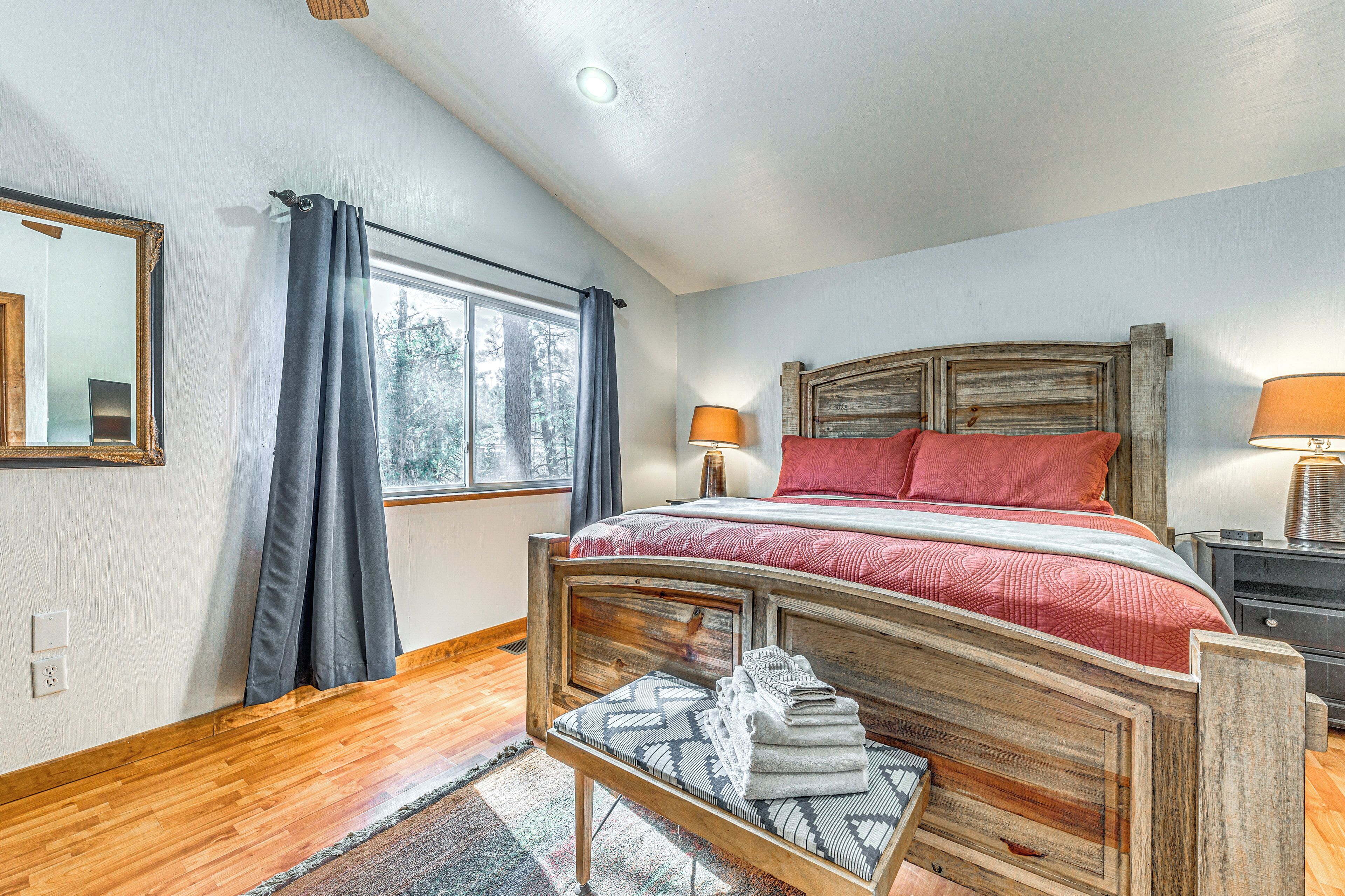 A bedroom with a wooden bed frame, a window with curtains, a bedside table with a lamp, and a mirror on the wall.