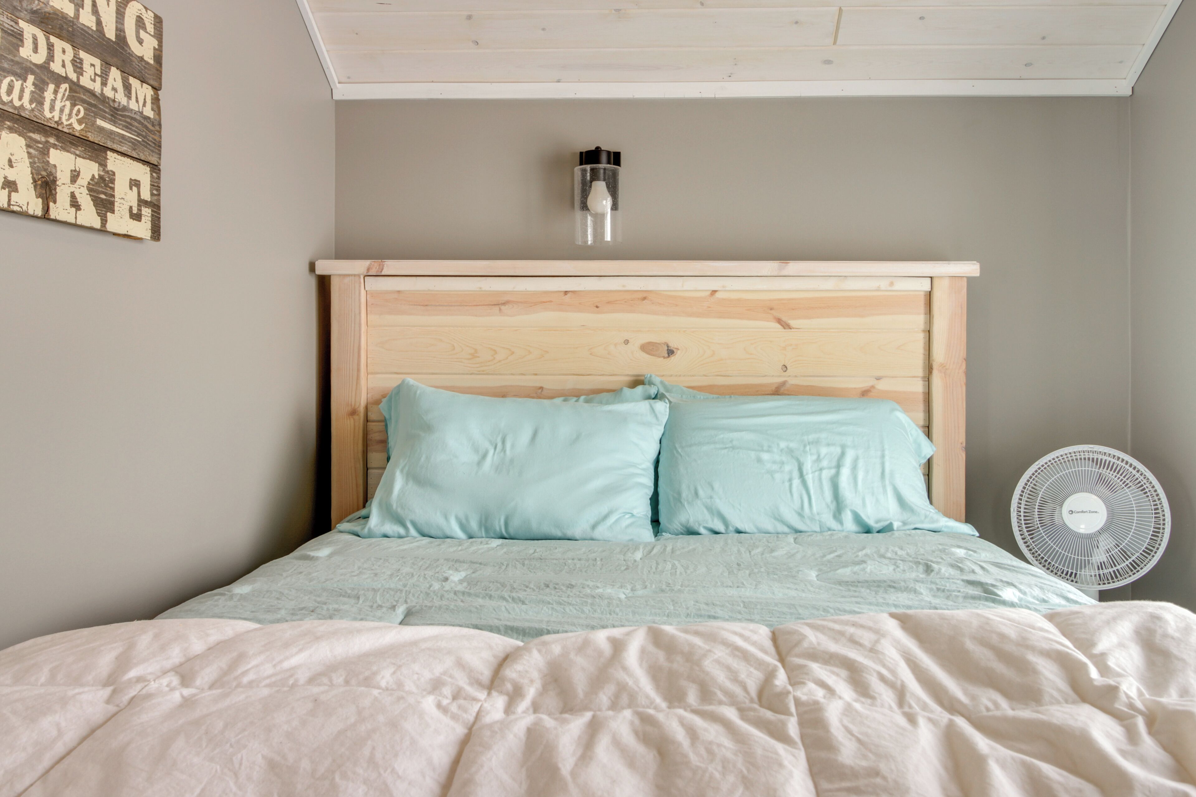 A bedroom with a wooden headboard, a bedside table with a lamp, and a wall-mounted fan.