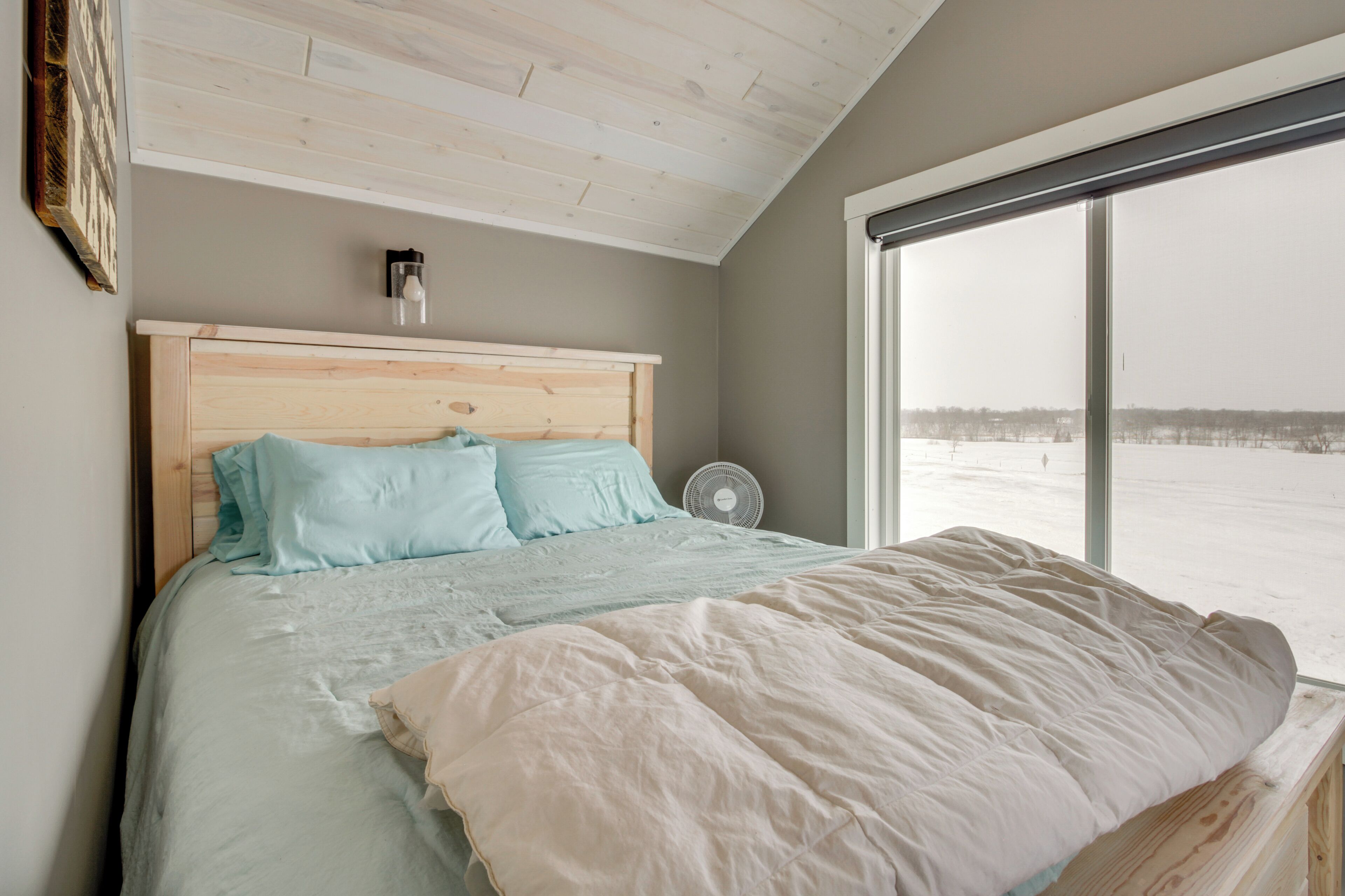 Ein Schlafzimmer mit einem hölzernen Kopfteil, einem großen Fenster mit Blick auf eine verschneite Landschaft und einem Nachttischventilator.