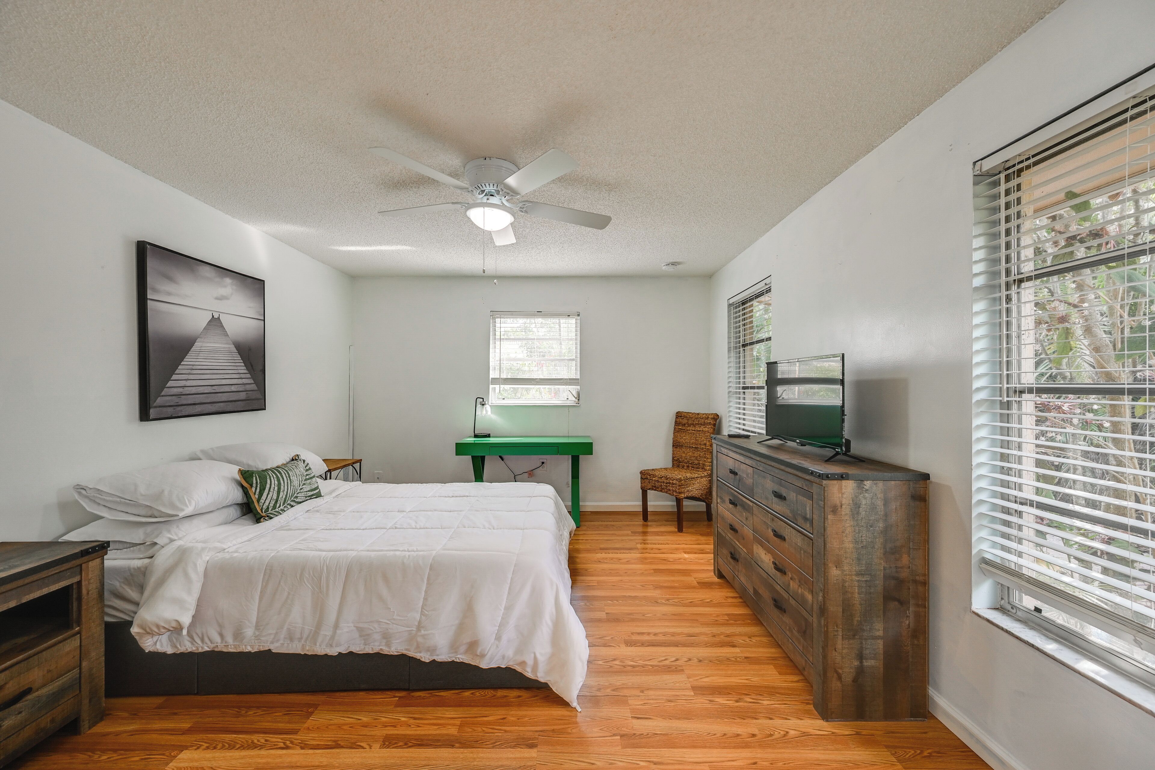 A bedroom with a bed, a desk, a chair, a dresser, a ceiling fan, and a window with blinds.