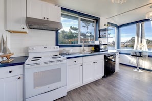 A modern kitchen with white cabinets, a stainless steel range hood, a double oven, and a dishwasher. There is a large window providing a view of the outdoors. - Fire Pit & River View: Serene Stay in Seaside! (Seaside)