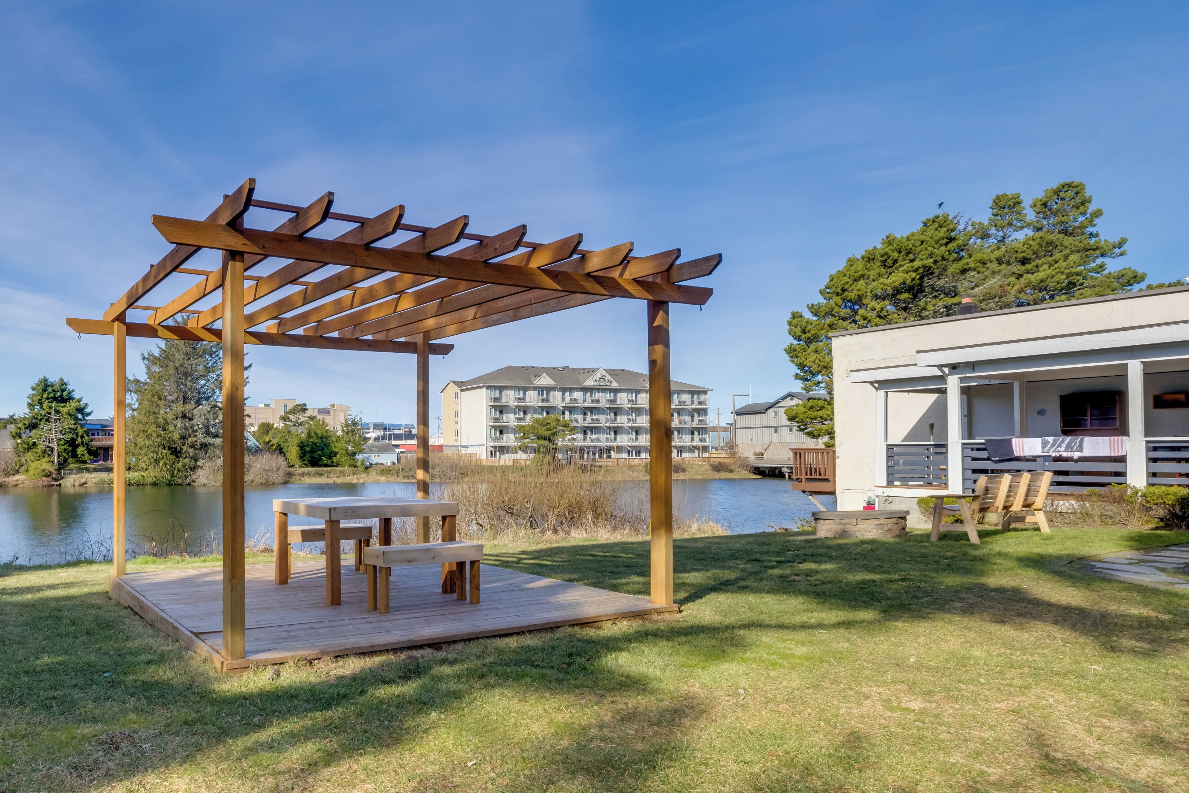 A wooden pergola with benches, a grassy area, a lake, and a building with a balcony in the background.