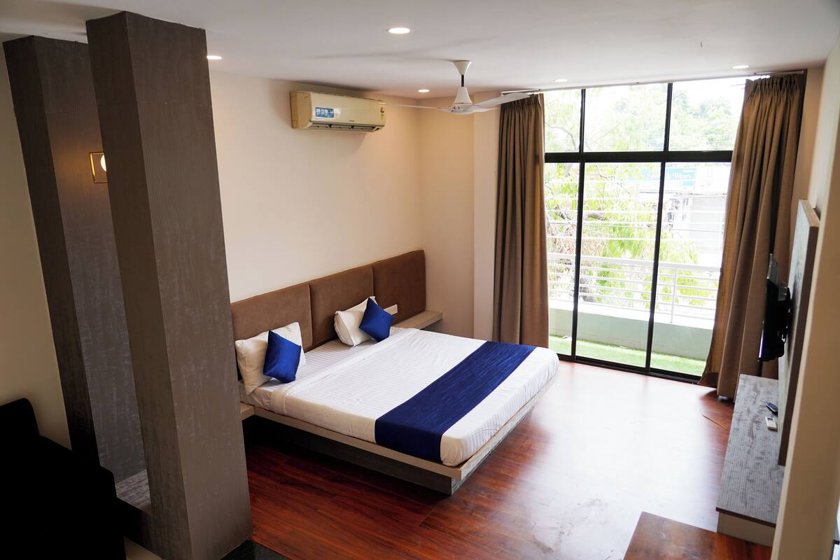 A modern hotel room with a large bed, a ceiling fan, an air conditioning unit, and a view of the outdoors through a large window.