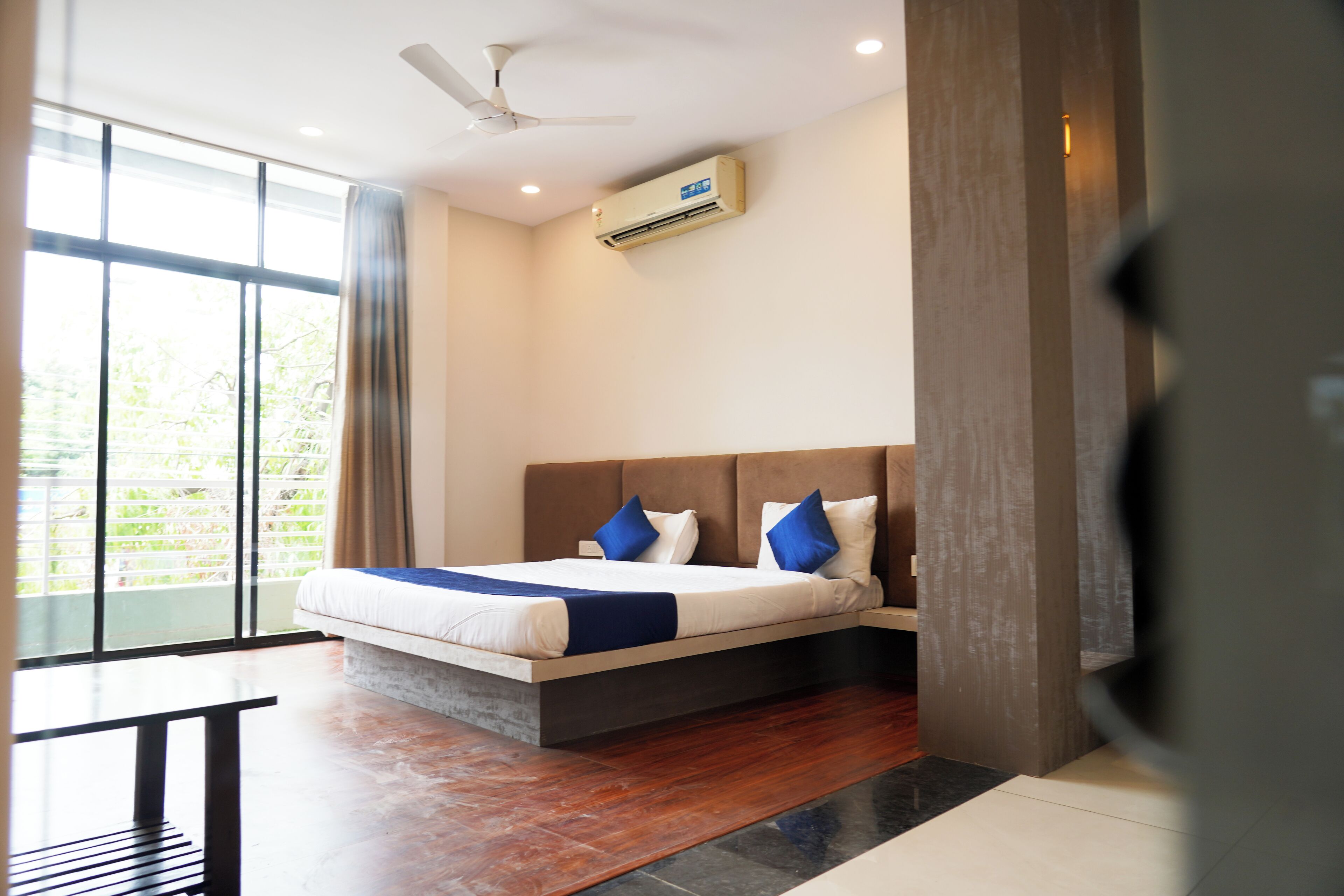A modern hotel room with a large bed, a ceiling fan, an air conditioning unit, and a view of greenery through the window.